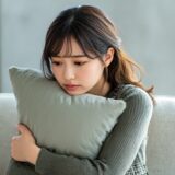 ソファでクッションを抱えながら落ち込む20代の女性